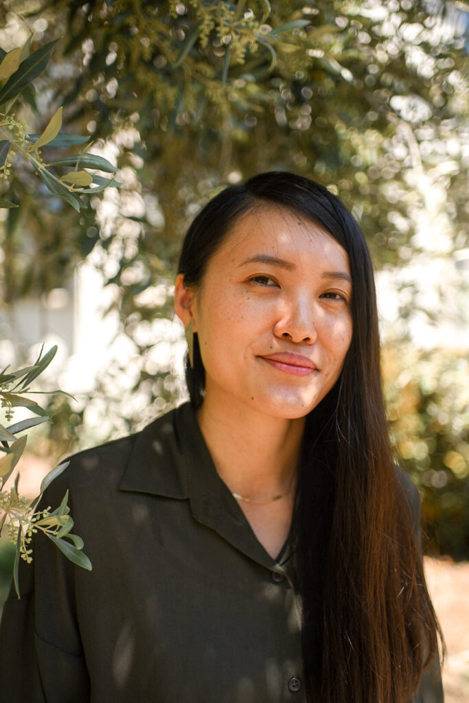 Headshot of Jade Cho from the shoulders up. She is smiling in a green button-up under a eucalyptus tree.